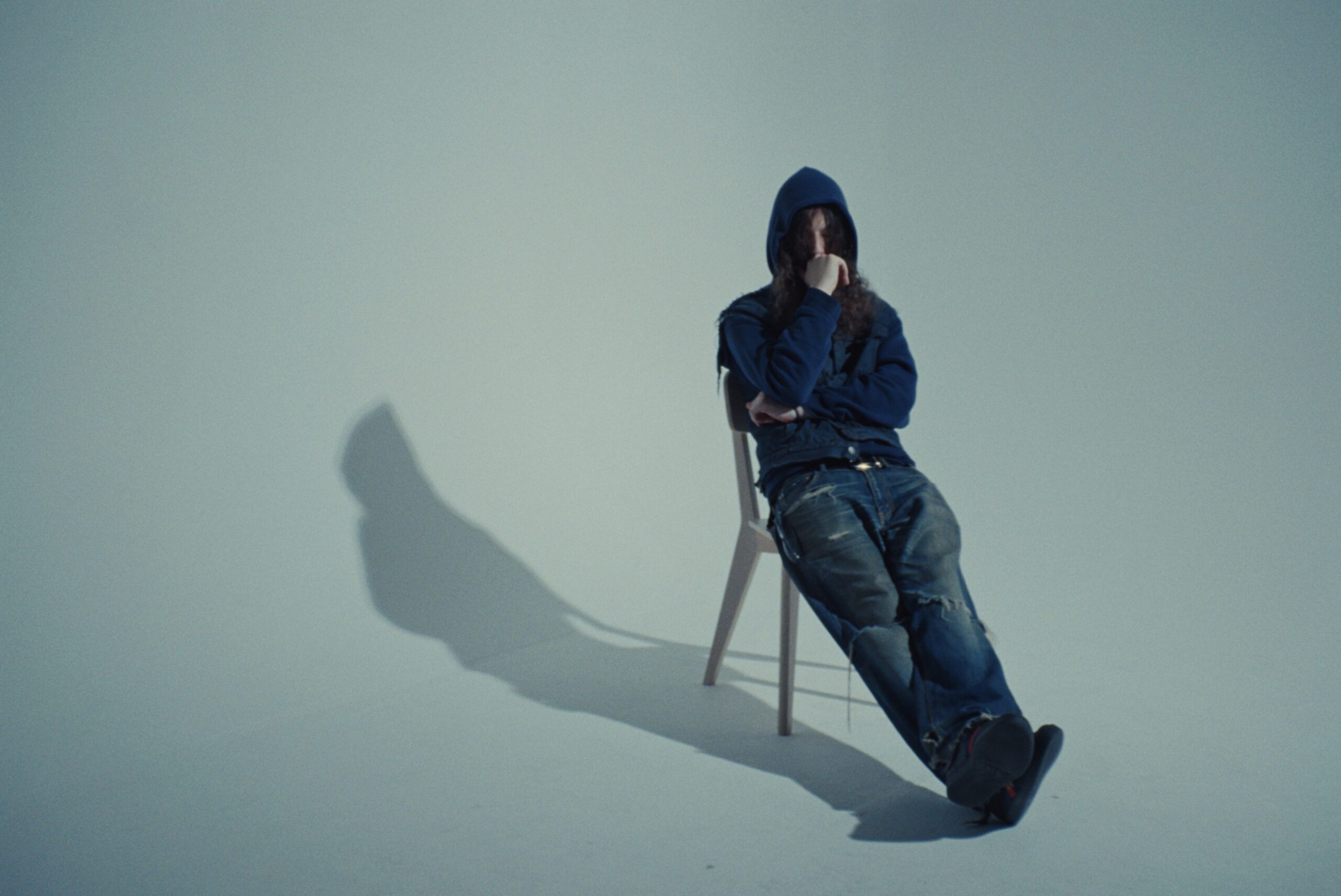 Person in a blue hoodie and jeans sits on a chair against a plain background. The individual is resting their chin on their hand, casting a shadow on the floor.